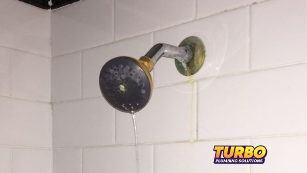 A showerhead with water leaking from the pipe connection against a tiled wall; visible water stains indicate ongoing plumbing issues and low water pressure. Turbo Plumbing Solutions logo in bottom right corner.