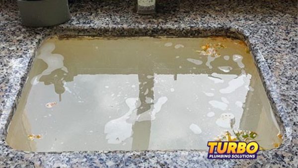 A kitchen sink filled with cloudy, dirty water and food debris signals drains blocking or blocked drains. The Turbo Plumbing Solutions logo is visible in the bottom right corner.