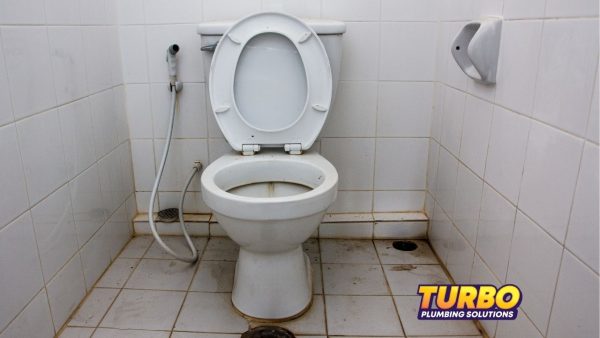 A white toilet with an open lid sits in a tiled, unused bathroom, showing visible dirt and stains on the floor and toilet. "Turbo Plumbing Solutions" logo is in the bottom right corner.
