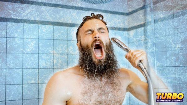 A bearded man reacts with discomfort while taking a handheld shower in a tiled bathroom, frustrated by hot water failure; "Turbo Plumbing Solutions" logo appears in the corner.