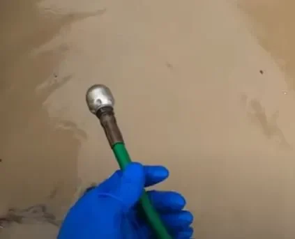 A gloved hand is holding a green-handled drain cleaning tool with a metal spherical end over a beige surface.