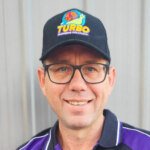A man wearing glasses and a black cap that reads "Turbo Plumbing Solutions." He is smiling and dressed in a purple and black top.