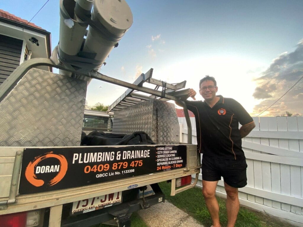 A smiling plumber standing beside his well-equipped work truck at dusk.