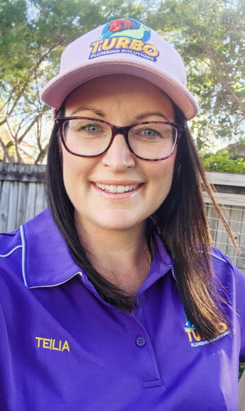 A woman wearing glasses, a pink hat, and a purple shirt that says "Teilra" smiles in an outdoor setting. The hat and shirt display the "Turbo Plumbing Solutions" logo.