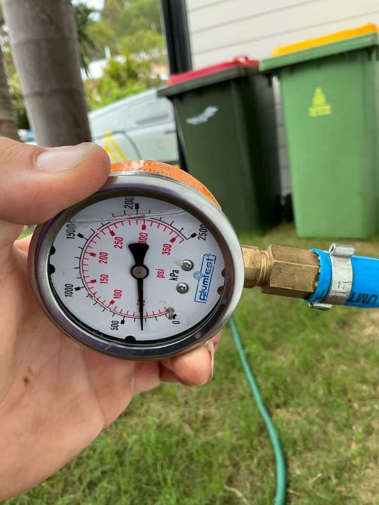 A hand holds a pressure gauge reading about 220 kPa, attached to a blue hose—part of a water pressure testing process—with bins and grass visible in the background.