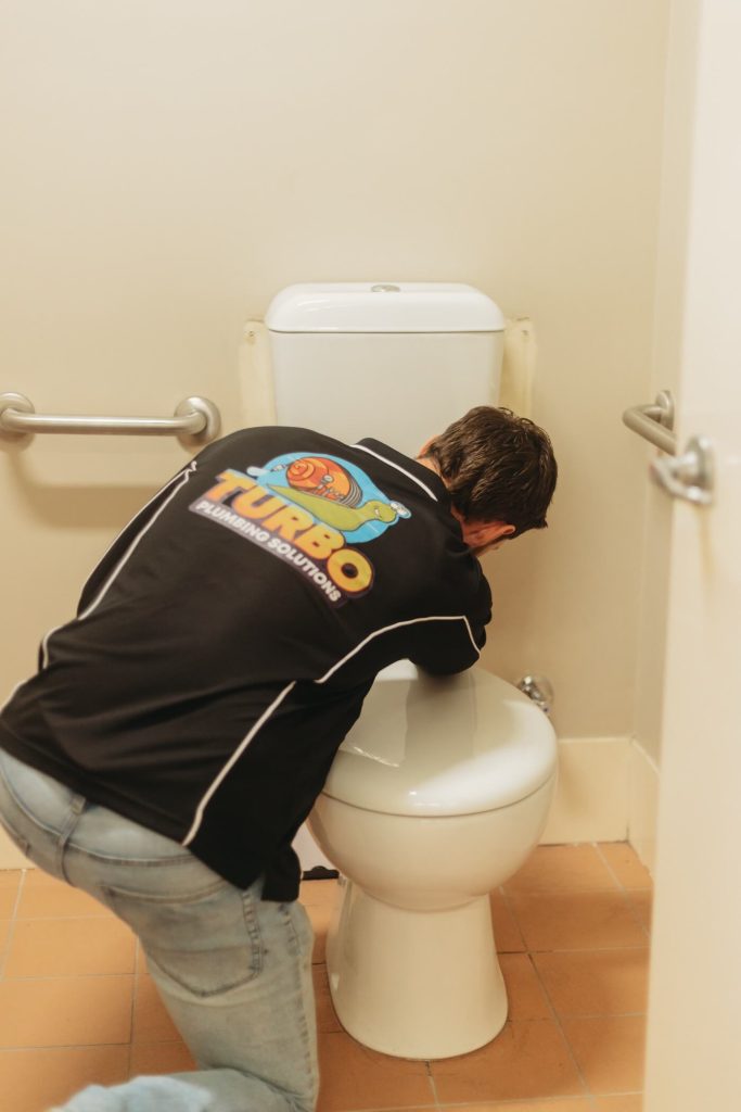 A person wearing a "Turbo Plumbing Solutions" shirt is kneeling and working on a toilet in a restroom with beige tiles and grab bars.