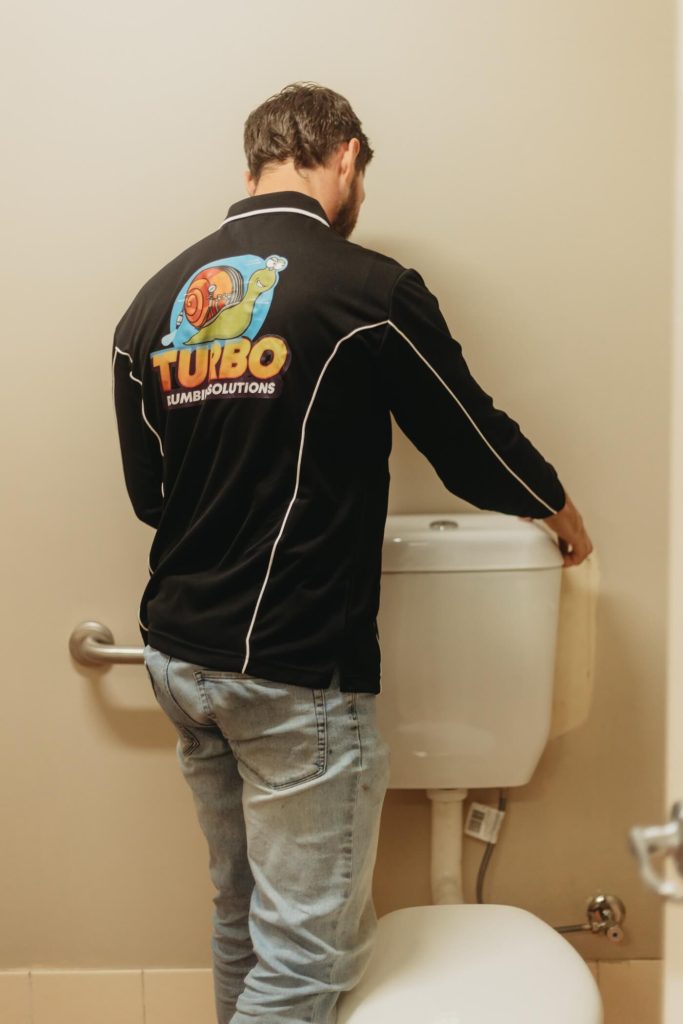 A plumber in a black "Turbo Plumbing Solutions" shirt is inspecting or repairing the tank of a white toilet in a restroom.