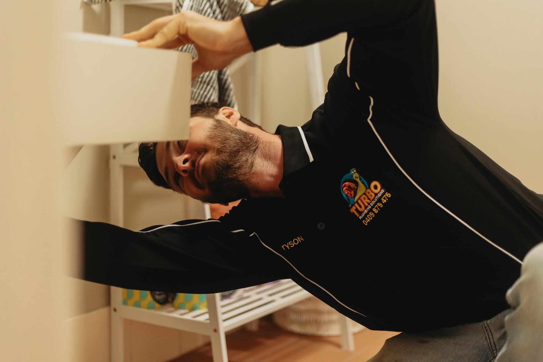 A man in a black shirt labeled "Turbo" inspects or repairs plumbing under a sink in a bathroom.