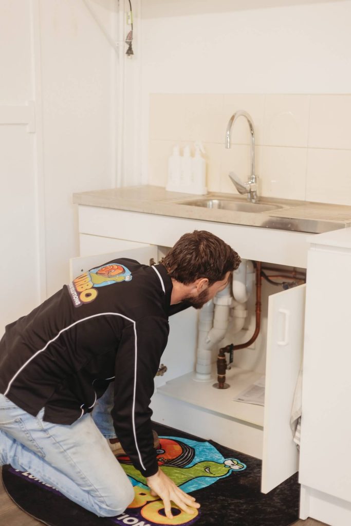 A man kneels on a mat and inspects the plumbing under a white sink in a modern kitchen or laundry area.