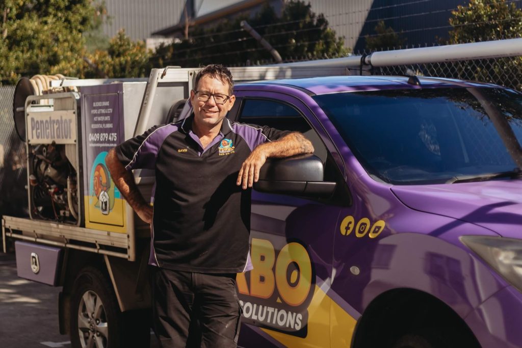 A man wearing glasses and a work uniform stands smiling with one arm resting on a purple service vehicle branded "FABO Solutions" in an outdoor setting.