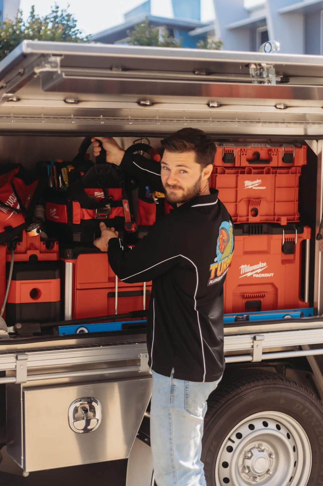A man in a black shirt and jeans stands beside an open truck compartment filled with organized red toolboxes and equipment.