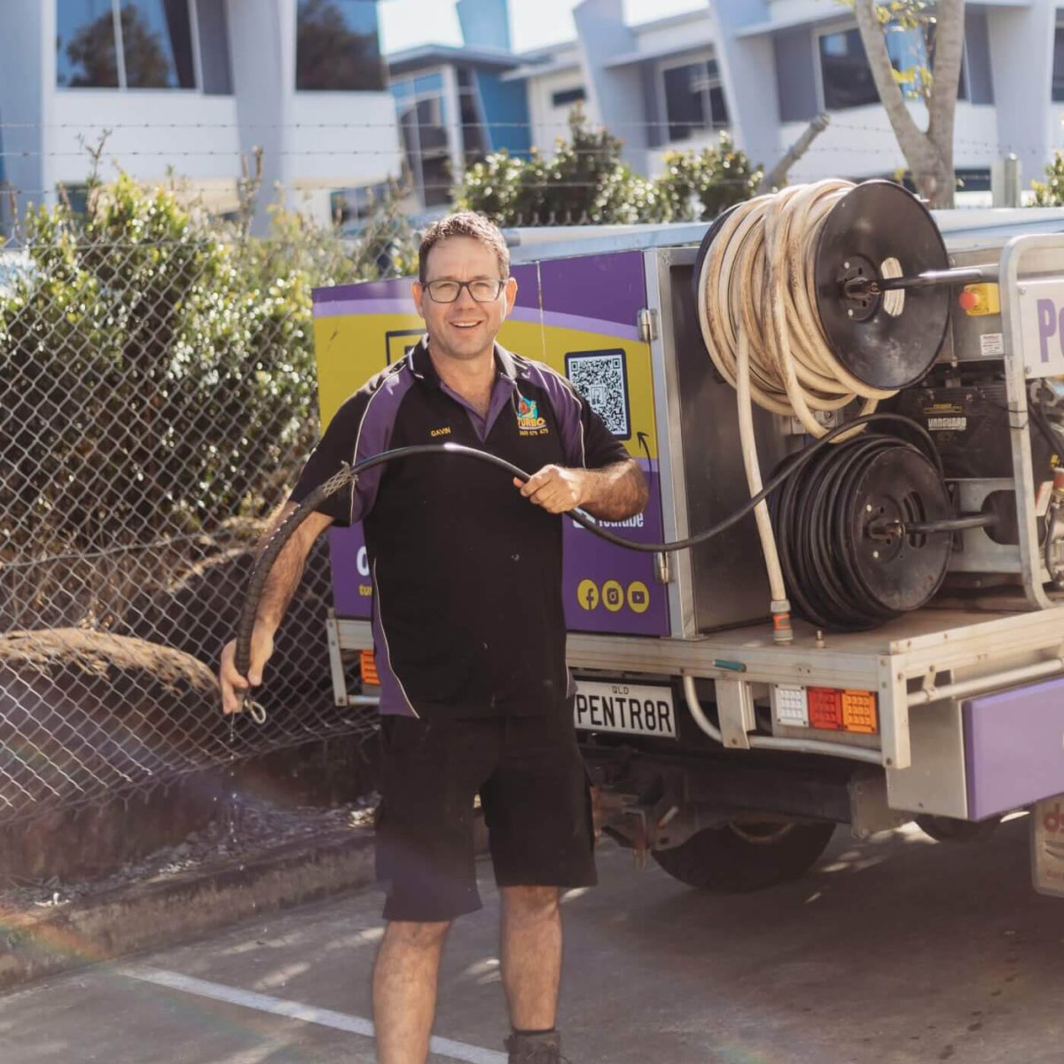 A man in a black uniform stands by a utility truck, holding a hose reel in a parking lot near a building.