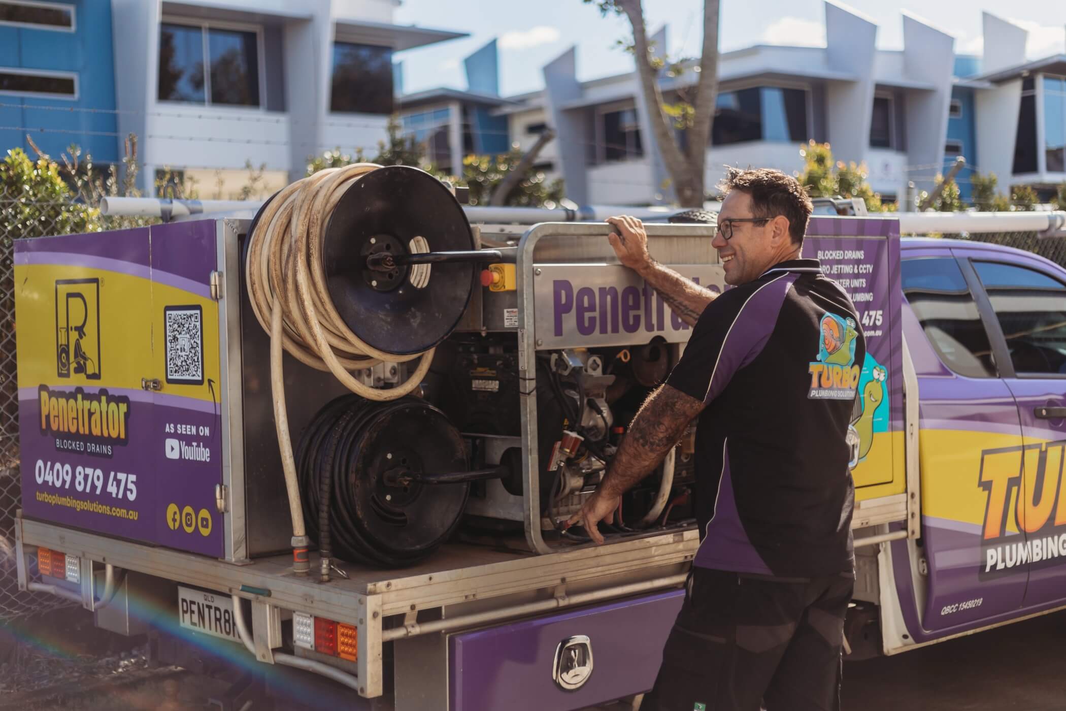 A plumber stands next to a work truck equipped with hoses and tools, parked outside modern buildings on a sunny day.