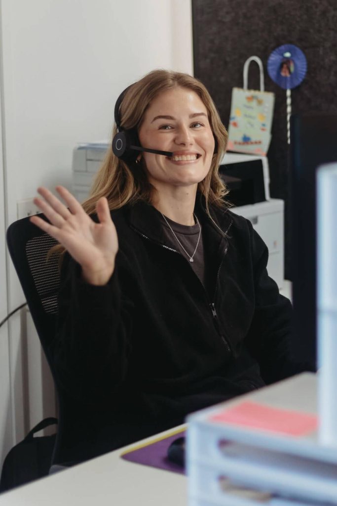 A woman wearing a headset sits at a desk and smiles while waving at the camera.