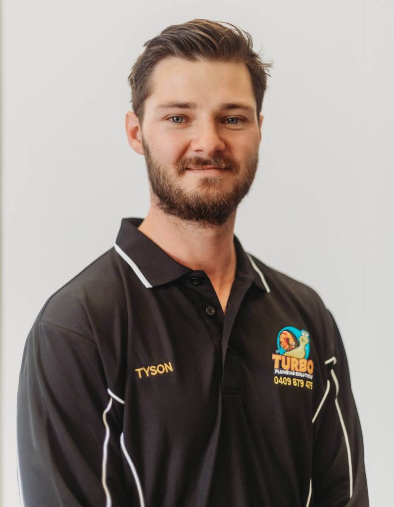 A man with short brown hair and a beard, wearing a black "Turbo Plumbing & Roofing" polo shirt with the name "Tyson" embroidered on it, stands against a plain white background.