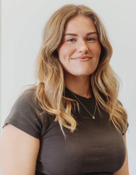 A woman with wavy light brown hair wearing a dark short-sleeve shirt and a necklace stands against a plain light background, smiling slightly.
