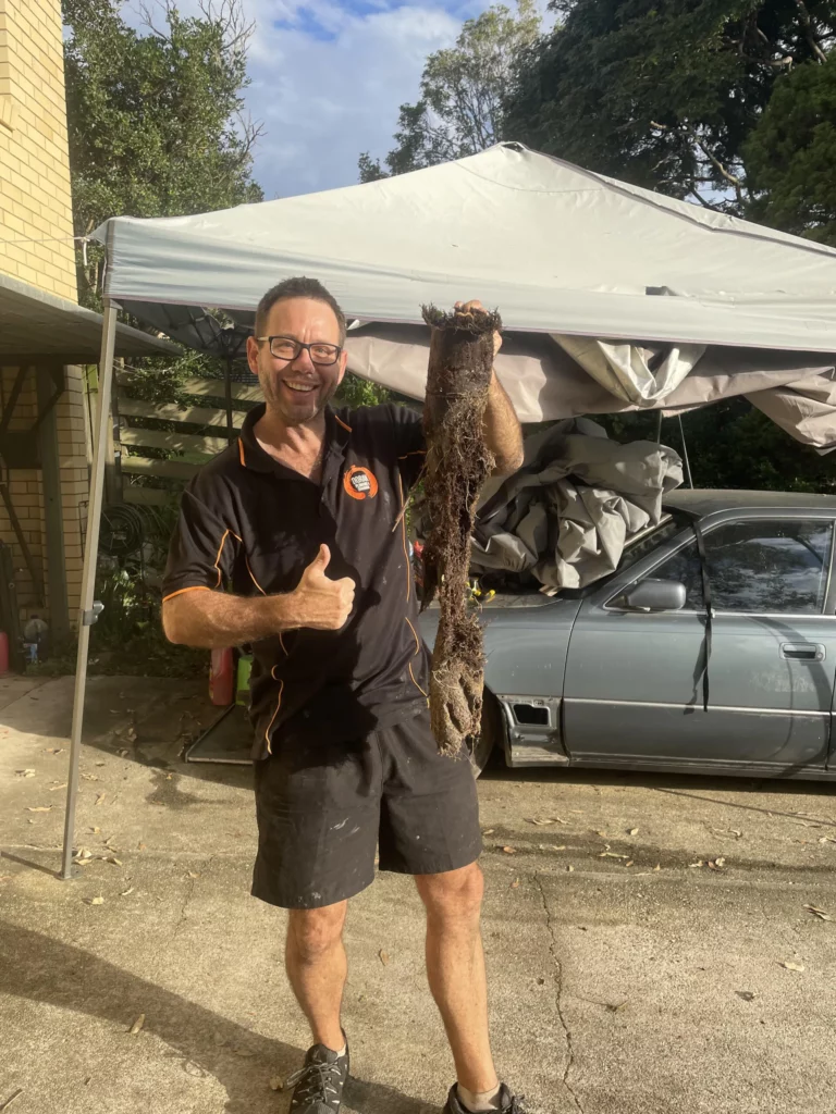A man in a black shirt and shorts holds a large, muddy tree root in one hand and gives a thumbs up with the other hand, standing in a driveway next to a silver car and a canopy.