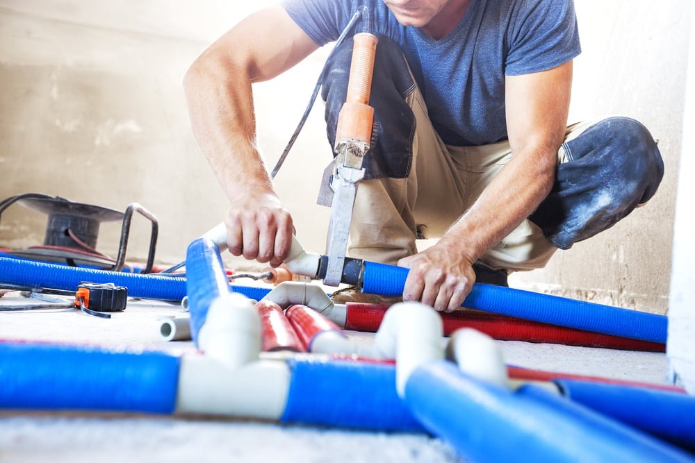 A person in a blue shirt and work pants, equipped with trade licenses, uses a welding tool to connect plastic pipes on a floor, surrounded by other piping components and tools.