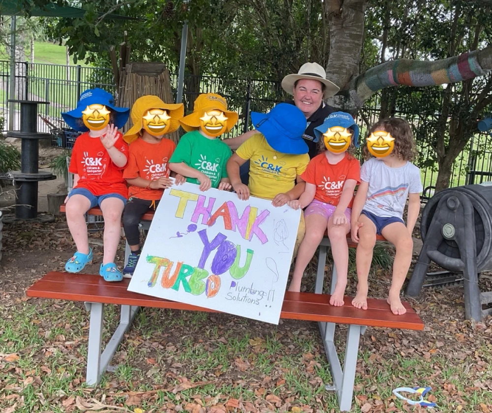 A group of children in colorful shirts and hats sit on a bench outdoors, holding a "Thank You" sign. An adult in a hat sits with them. Trees and a fence are visible in the background.