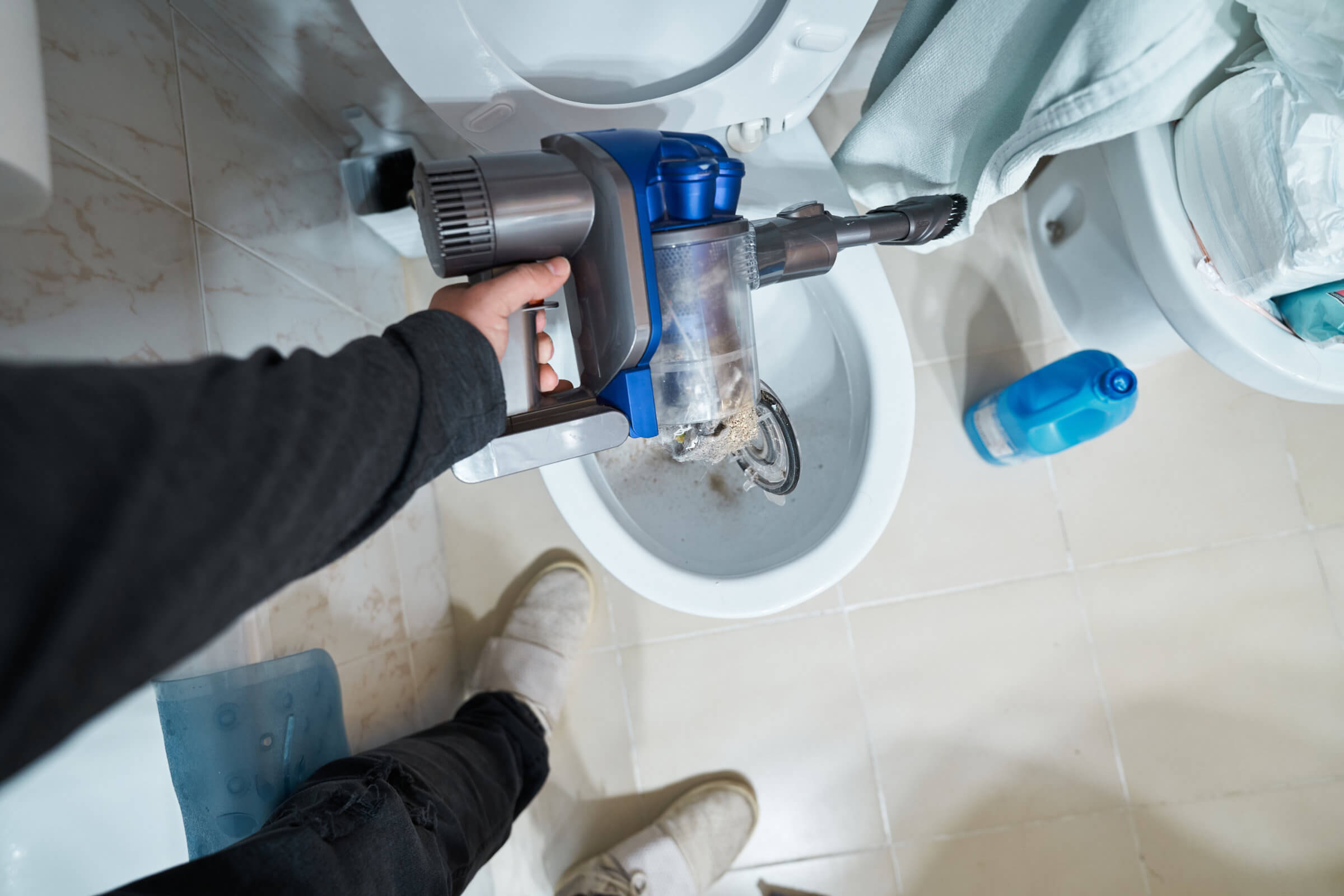 Person emptying contents of a vacuum cleaner into a toilet in a bathroom, likely contributing to a blocked drain that may require drain repairs.