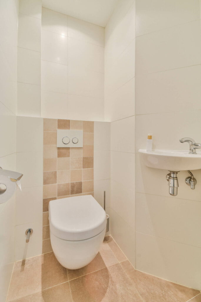 A small bathroom with a wall-mounted toilet, beige tiled walls, a compact sink with a faucet, and a toilet paper holder. Ensure regular maintenance to avoid blocked drains or needing drain repairs in such tight spaces.