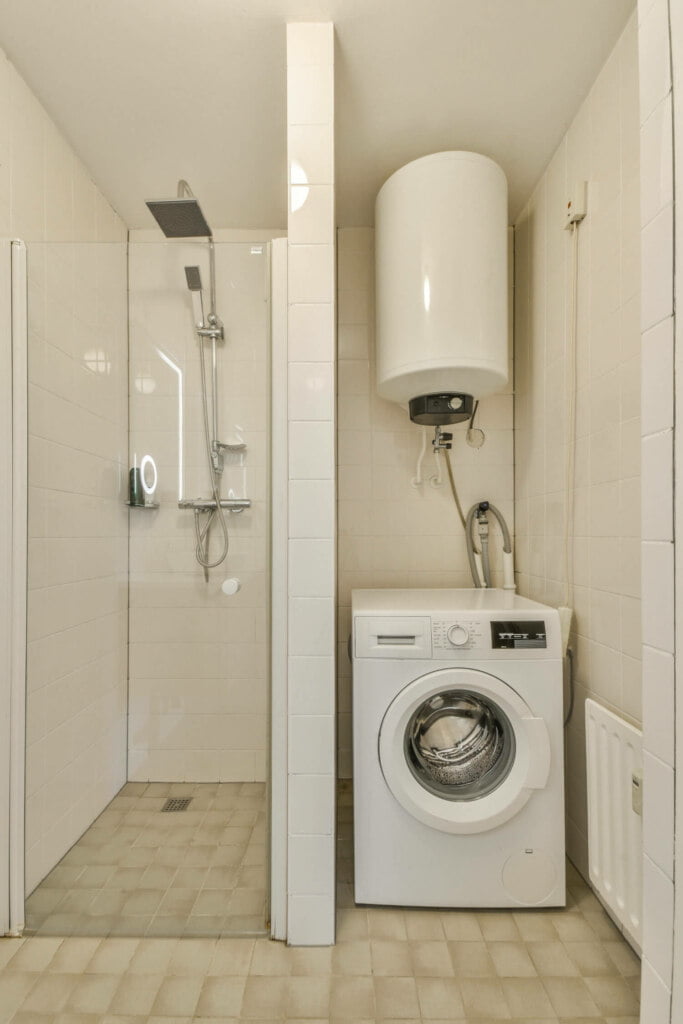 A small bathroom features a shower area with a glass partition, a wall-mounted water heater, and a front-loading washing machine. The floor is tiled with square beige tiles, and blocked drain repairs have ensured smooth water flow.