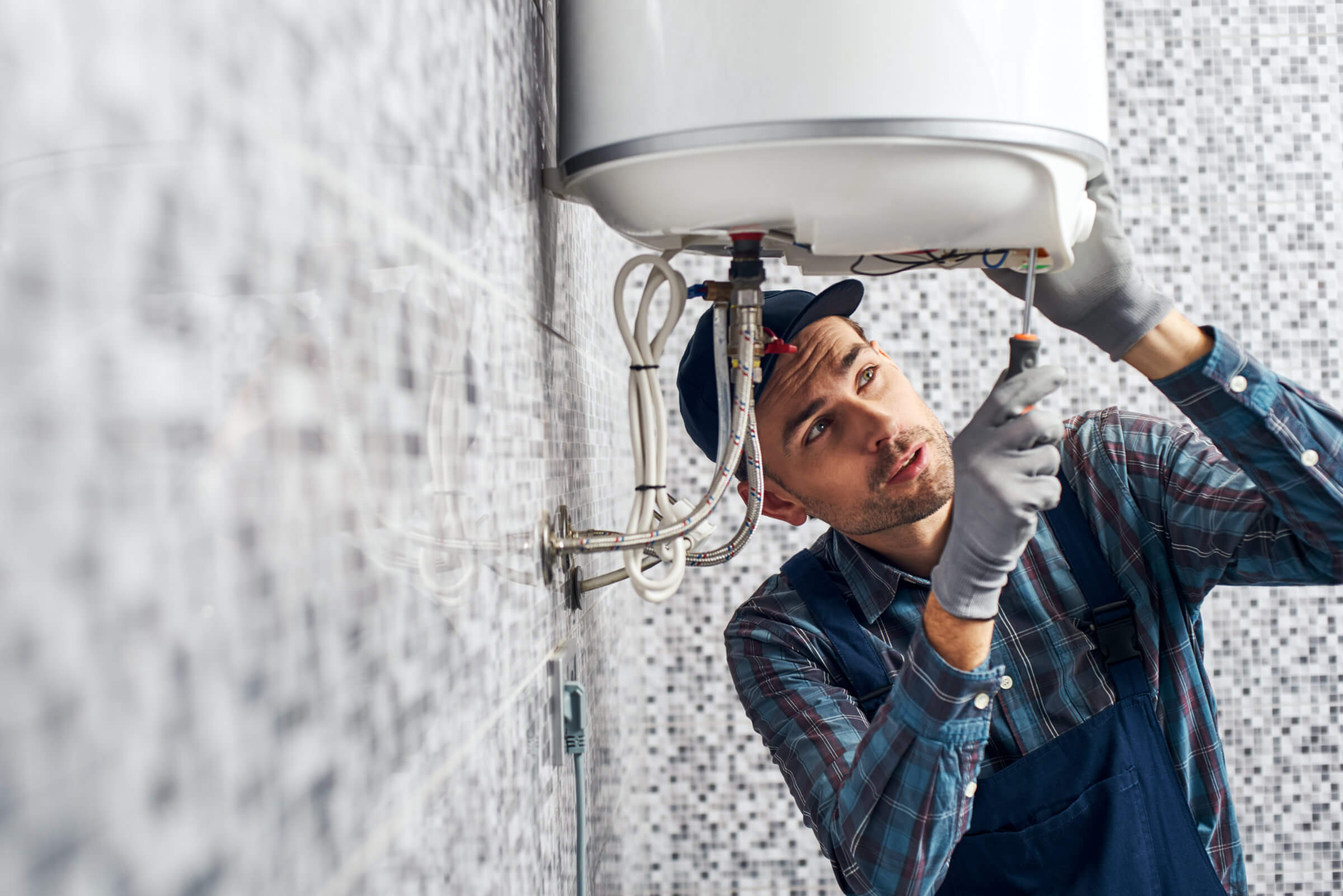 A man wearing a cap, checkered shirt, and overalls is using a screwdriver to repair a wall-mounted water heater in a tiled room, while also attending to a blocked drain nearby.