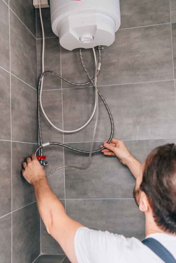 A person is adjusting the pipes connected to a wall-mounted water heater in a tiled room, addressing potential issues related to blocked drain repairs.