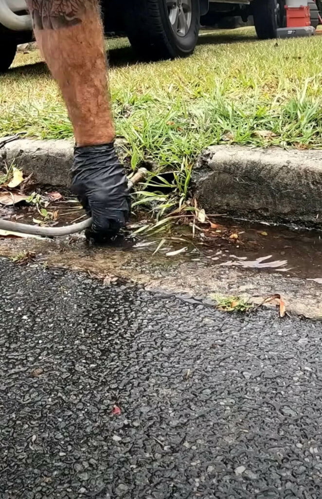 A person wearing a black glove uses a hose to clear debris from a blocked drain by the curb on a wet road, ensuring proper drain repairs.
