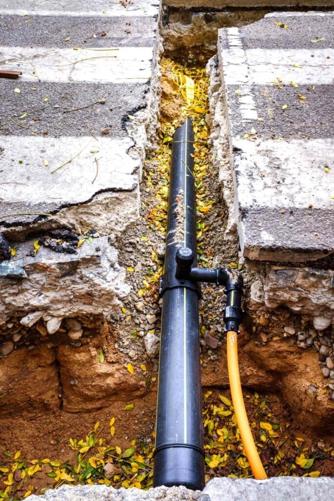 An exposed black pipe and yellow cable lie in a trench cut through a concrete sidewalk, with scattered yellow leaves around the trench—a clear sign of ongoing drain repairs.