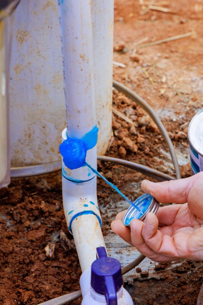 A person applies sealant tape to a white PVC pipe joint outdoors, with a dirt ground and other piping equipment visible in the background, ensuring the blocked drain is properly fixed.