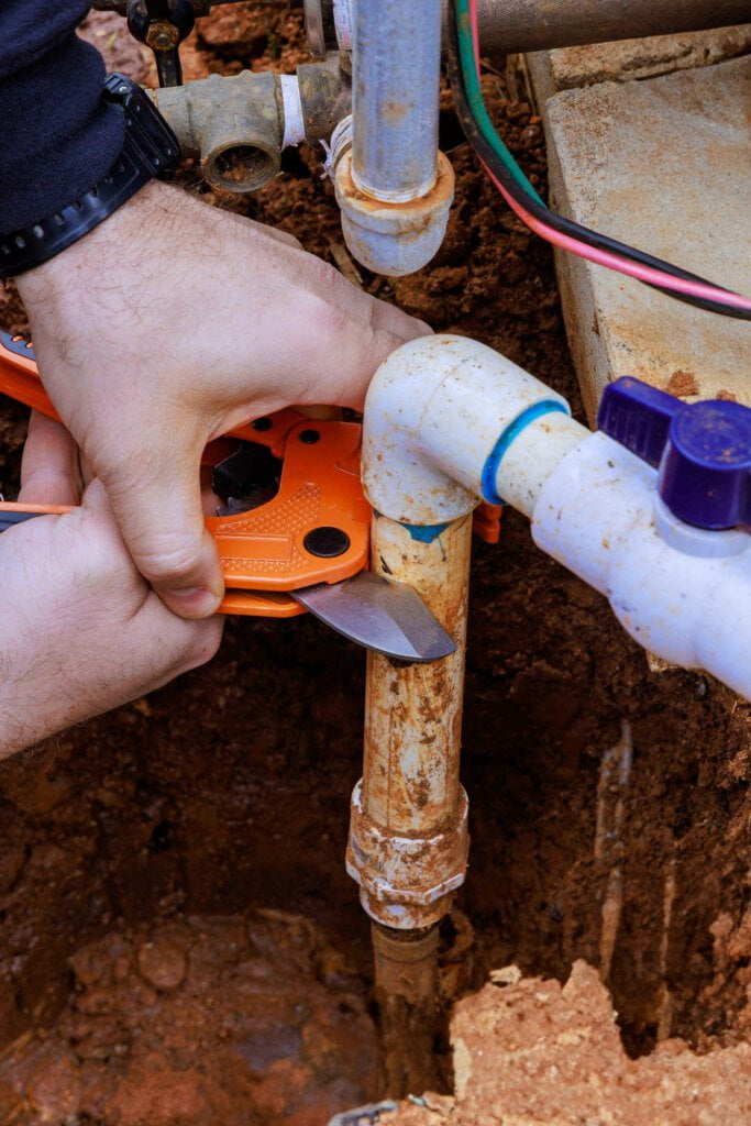 A person using an orange pipe cutter tool to perform blocked drain repairs, cutting through a rusted metal pipe in a muddy outdoor setting with various pipes and fittings strewn about.