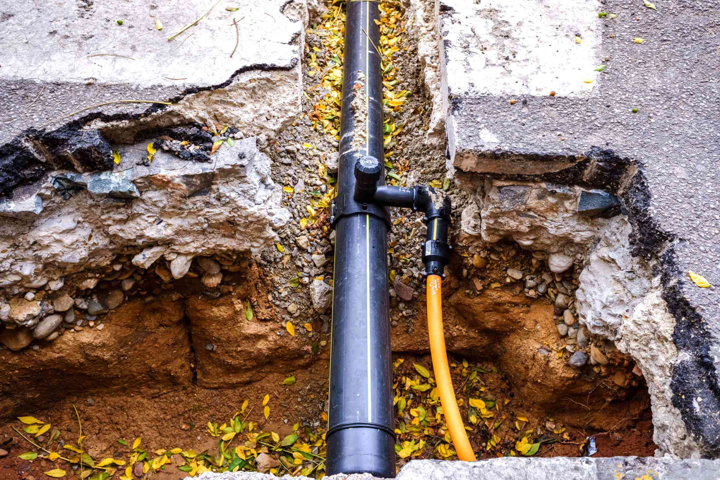 Exposed underground utilities, including a black pipe and an orange cable, run through a trench dug into the pavement. The area is surrounded by broken concrete and scattered leaves, often necessitating blocked drain repairs to prevent further damage.
