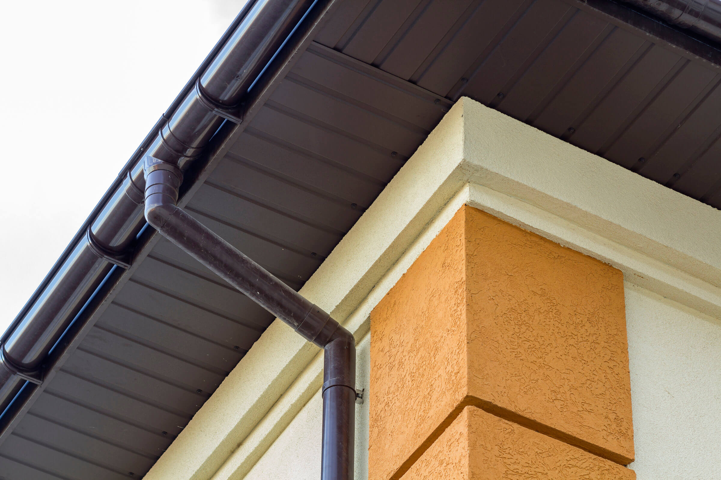 Close-up of a house corner with brown gutters and downspouts attached to a beige and orange external wall under a dark brown roof, seamlessly blending aesthetics with functionality, ensuring no need for frequent blocked drain repairs.