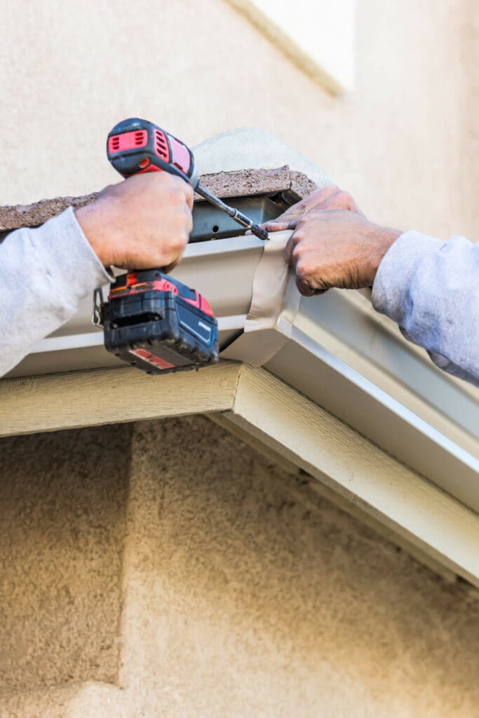 Two people are installing or repairing a rain gutter on a house, with one person using a drill and the other holding the gutter in place, ensuring effective drain repairs to prevent future issues.