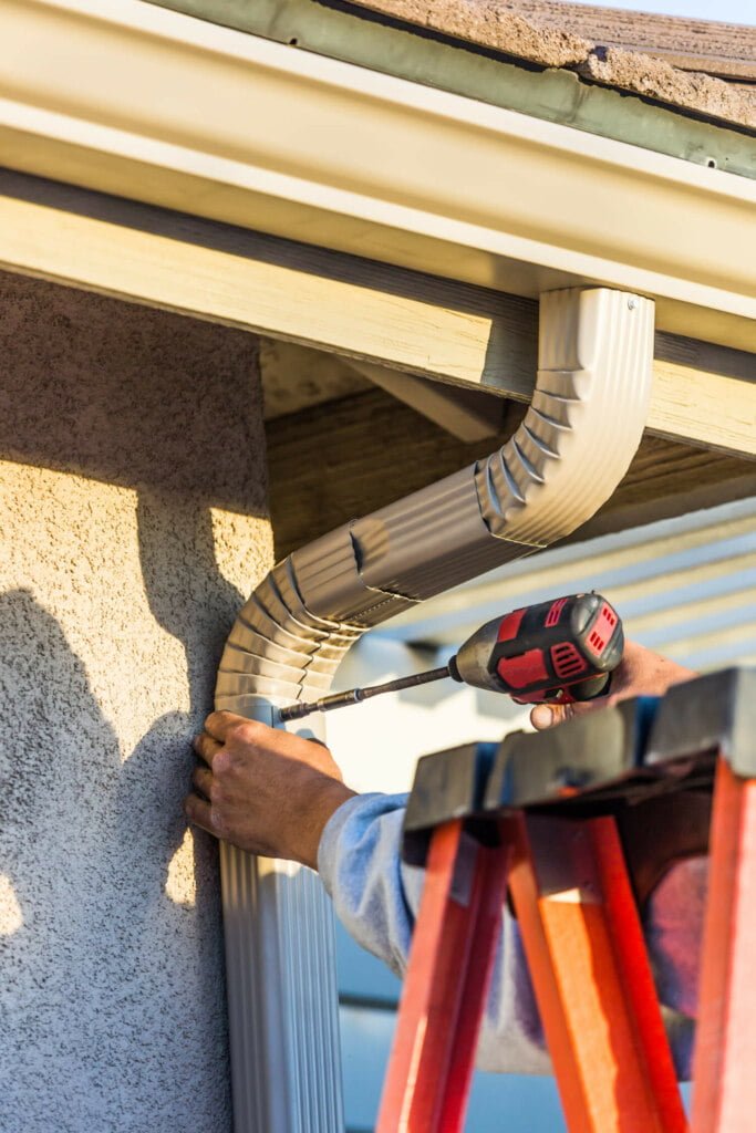 A person standing on an orange ladder uses a drill to secure an aluminum downspout to the side of a house near the roof, addressing blocked drain repairs.