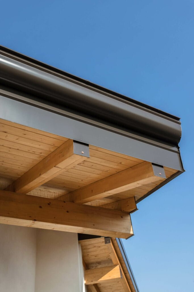Close-up view of the underside of a modern roof with exposed wooden beams and a metal gutter system against a clear blue sky, showcasing an impeccable home exterior that eliminates the need for blocked drain repairs.