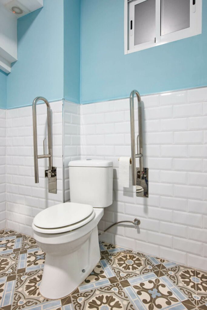 A bathroom with a white toilet, blue walls, white tiled wainscoting, patterned floor tiles, and two stainless steel handrails on either side of the toilet. A small window is above the toilet. Ideal for those needing accessibility while ensuring blocked drain repairs are easily managed.