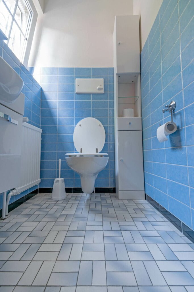 A bathroom with blue tiled walls features a wall-mounted toilet, a toilet brush, a toilet roll holder with a roll of paper, and a tall white cabinet with open and closed shelves. For any blocked drain issues, prompt drain repairs ensure everything remains in top condition.