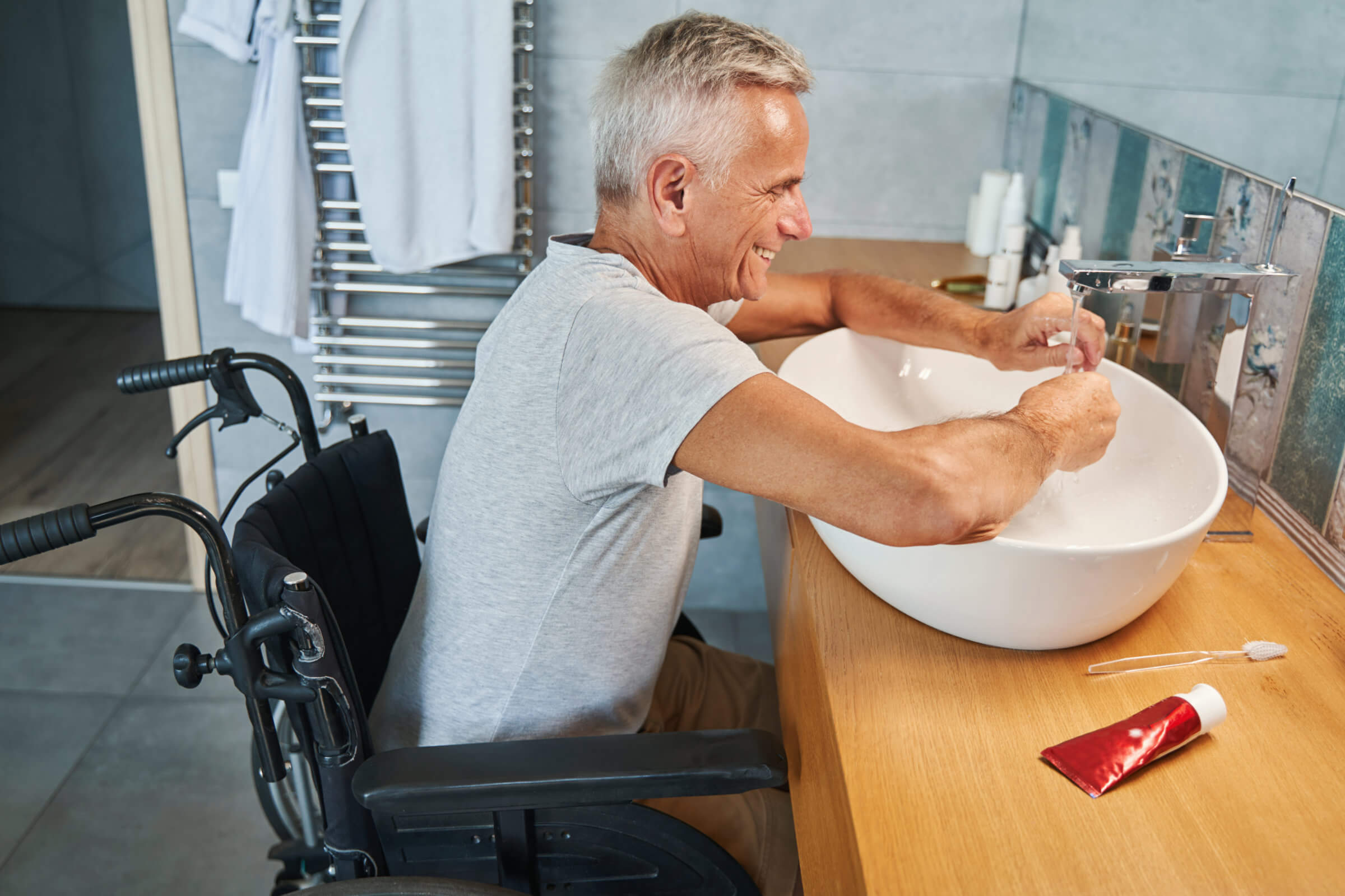 Elderly man in a wheelchair washing his hands at a bathroom sink with a smile, ensuring everything is clean and functional, even if it means addressing minor concerns like blocked drain repairs.