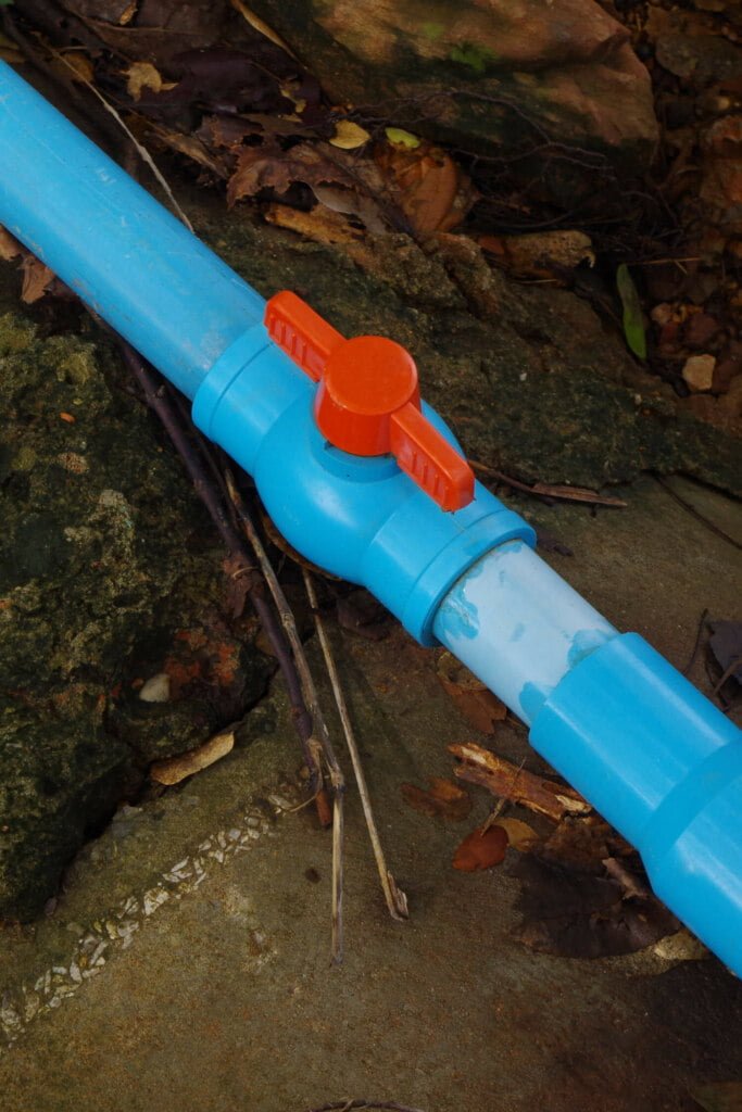 A blue plastic pipe with an orange valve handle is positioned on the ground, surrounded by rocks and dried leaves, ready for blocked drain repairs.