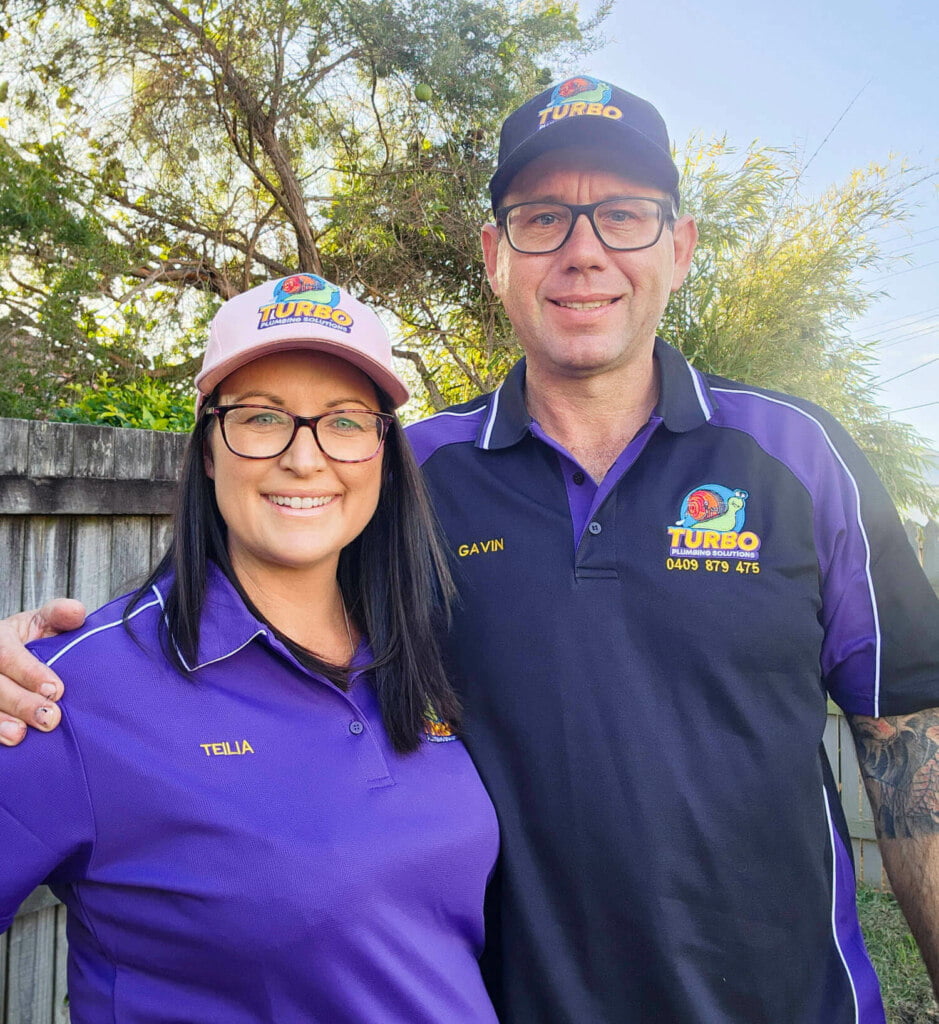Two people in matching uniforms, hats, and glasses, identified as Telia and Gavin, stand outdoors near a wooden fence, smiling at the camera. They both wear purple polo shirts with a company logo and names.