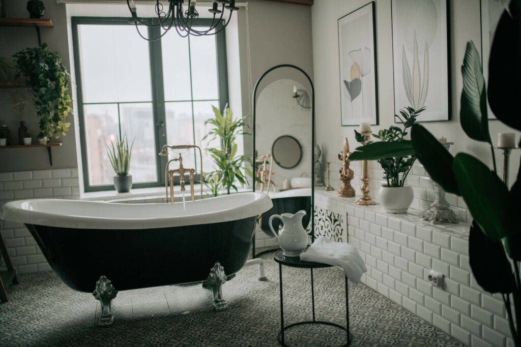 A modern bathroom featuring a freestanding black and white bathtub, a large window, potted plants, framed wall art, and a small table with a white jug and folded towel showcases thoughtful bathroom modifications.