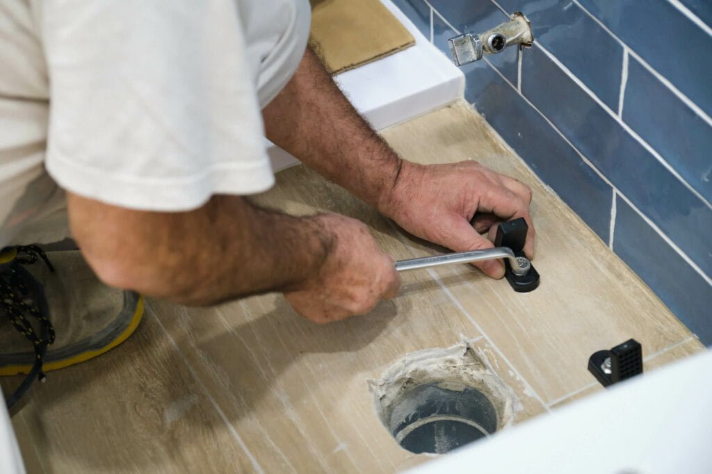 A person installs bathroom hardware on a wooden floor next to a hole, using a wrench. The wall features blue tiles, creating an ideal backdrop for the necessary blocked drain repairs.