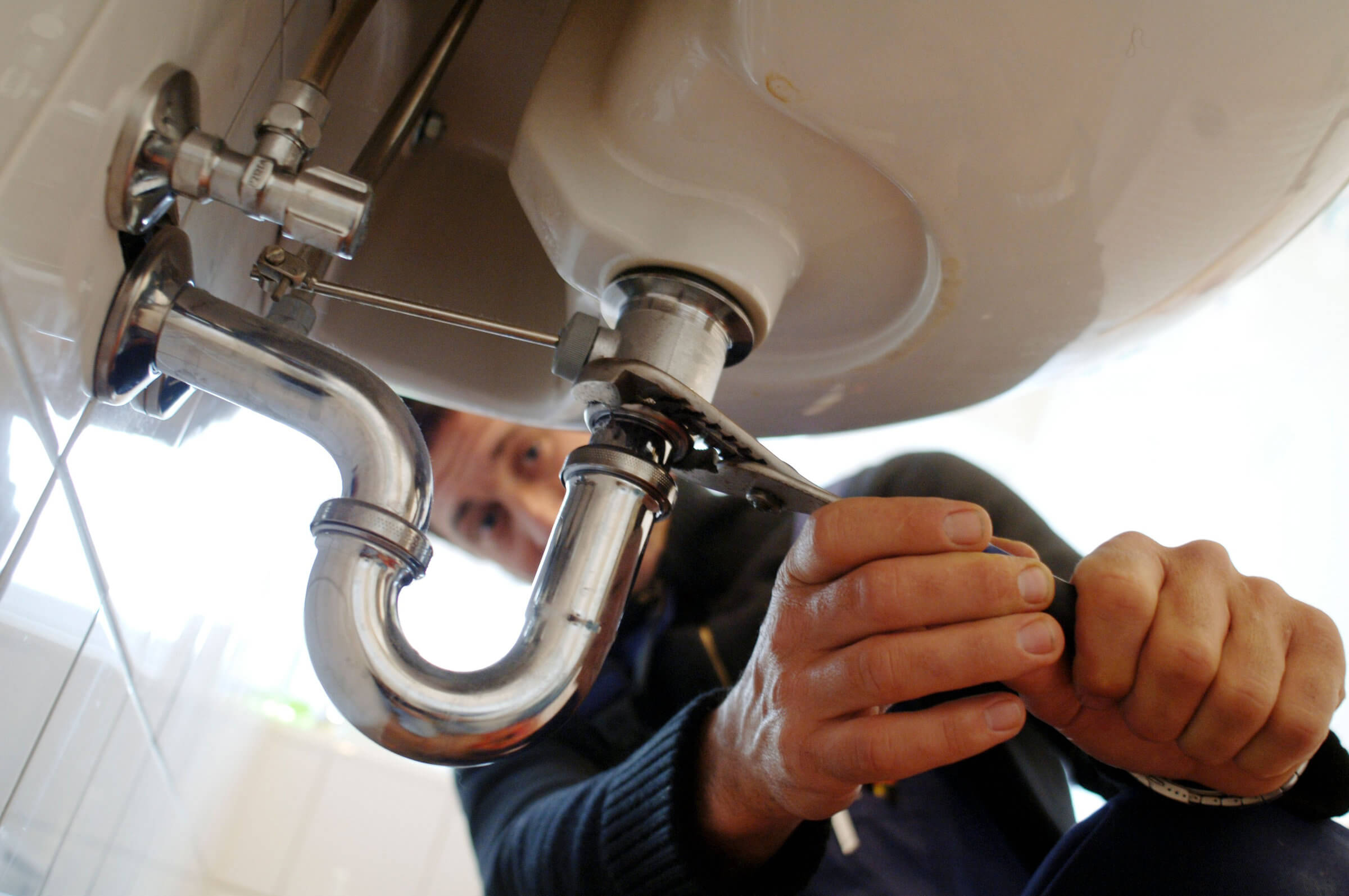 A plumber uses a wrench to tighten a pipe fitting underneath a sink, ensuring everything is secure while also checking for any potential blocked drain repairs that might be needed.