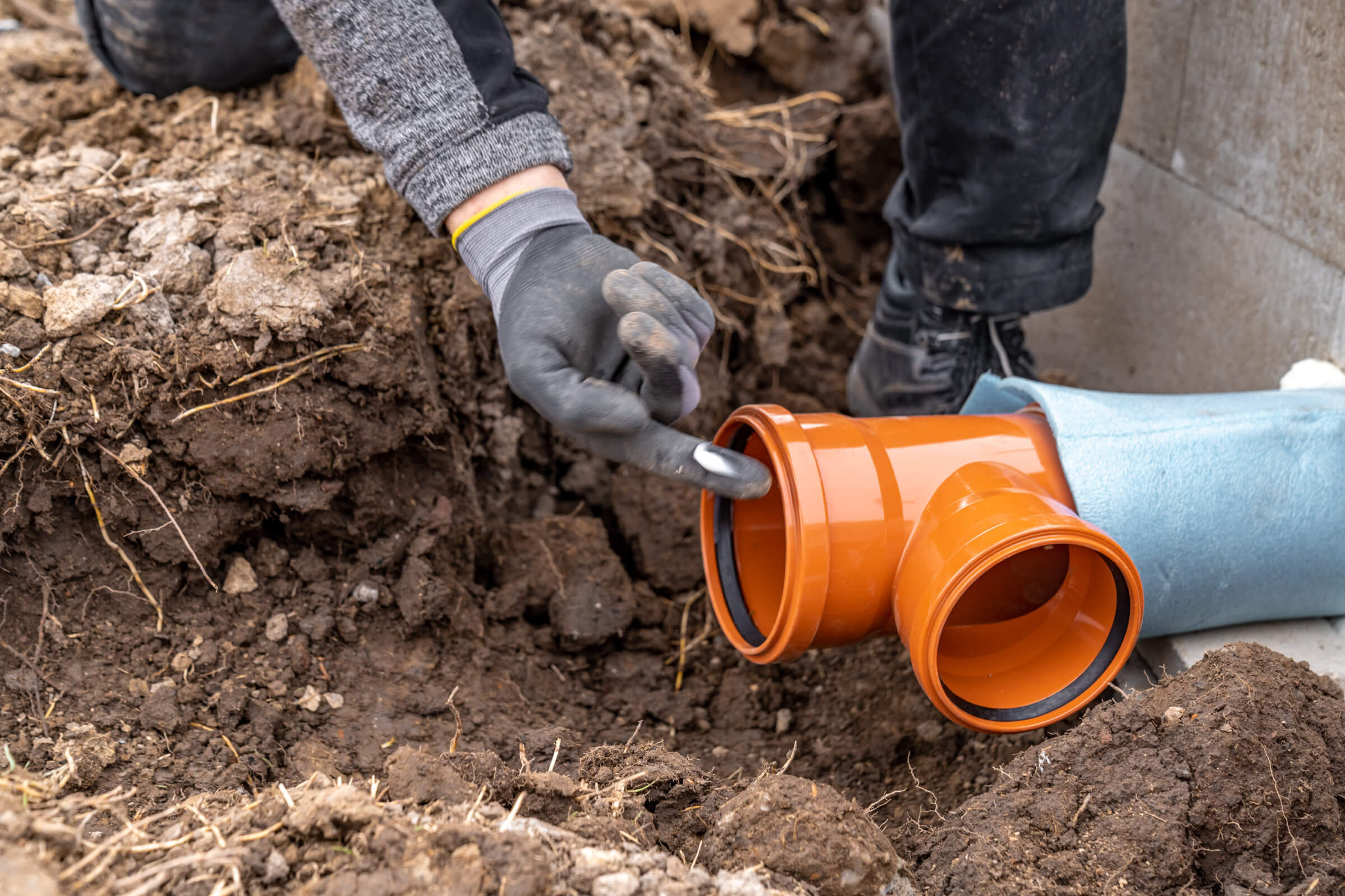 A person wearing gloves is handling a bright orange PVC pipe fitting in a muddy construction trench, working diligently to address a blocked drain. Another partially buried pipe is connected to the fitting, showcasing ongoing drain repairs in the challenging conditions.