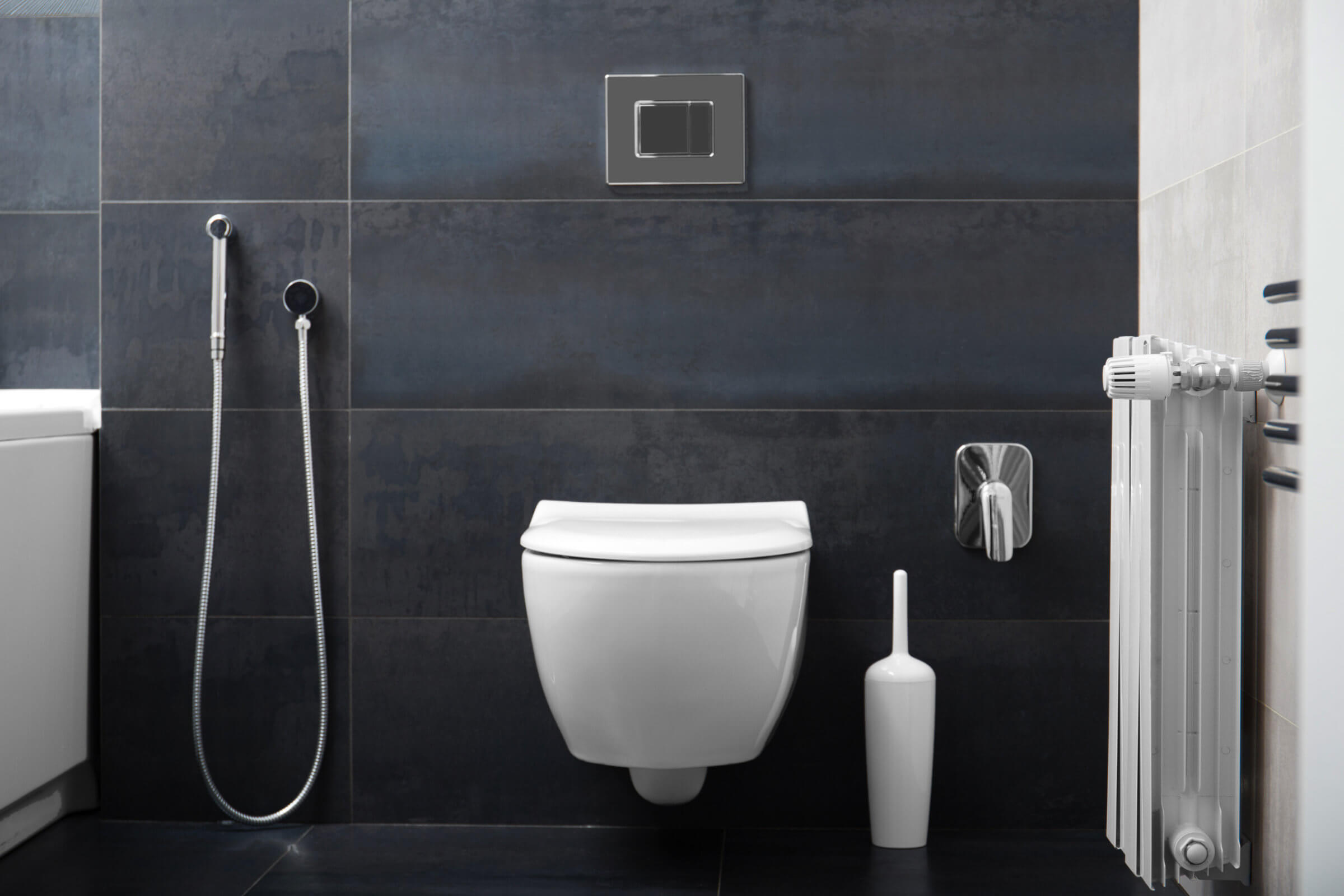A modern bathroom with black tiled walls, a wall-mounted toilet, a white toilet brush holder, a bidet spray, and a radiator on the right. The design ensures easy access for any blocked drain repairs that might be needed.