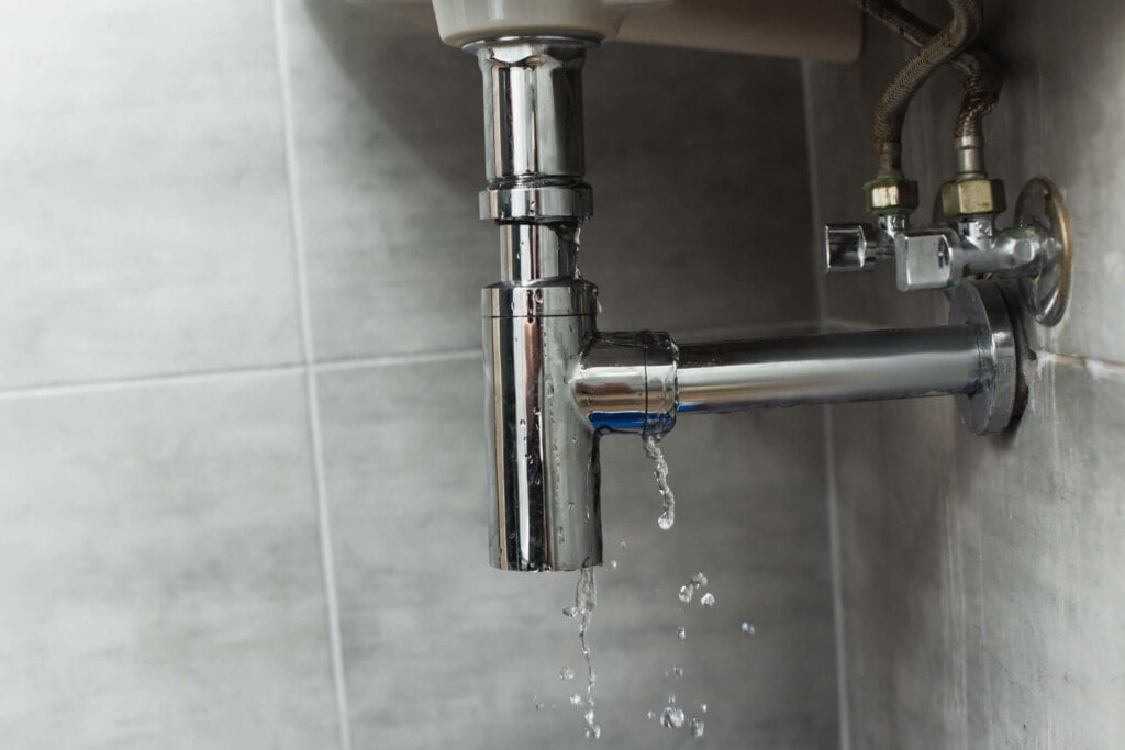 A leaking chrome pipe under a sink with water dripping into a tiled area may indicate the need for immediate Blocked Drain Repairs.