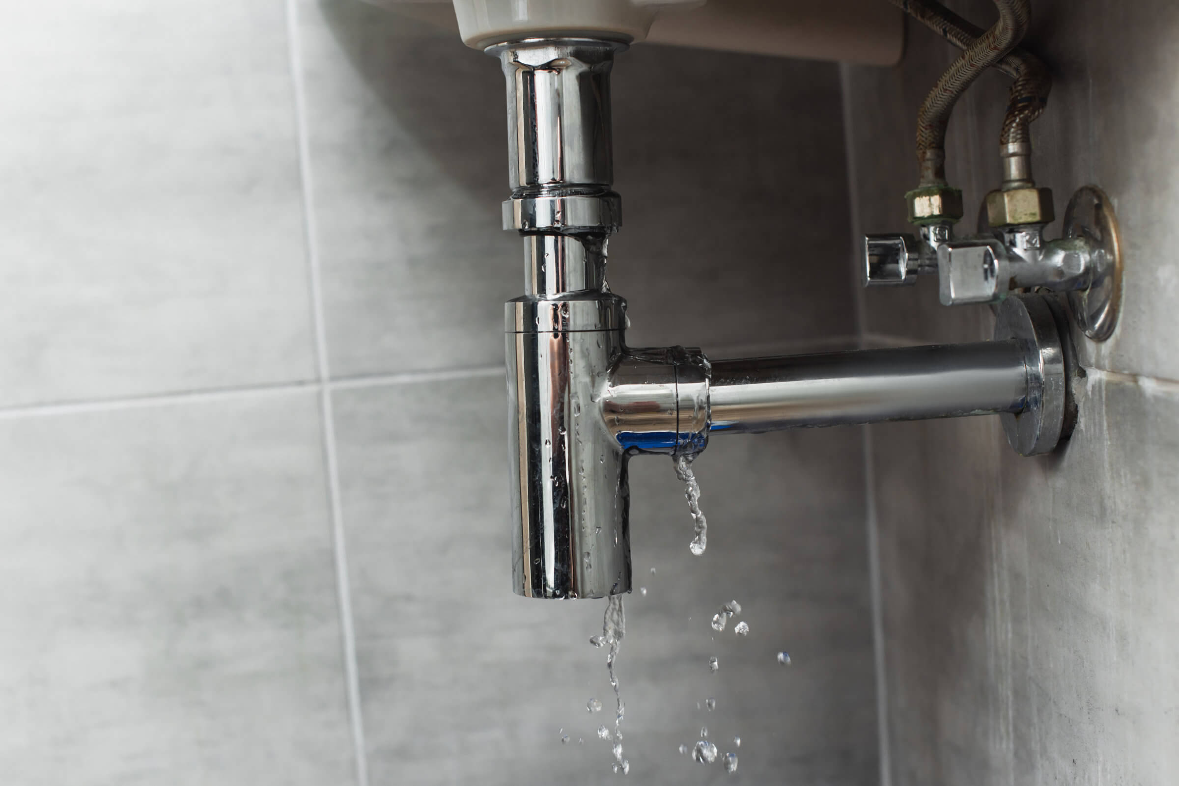 A leaking chrome pipe under a sink with water dripping into a tiled area may indicate the need for immediate Blocked Drain Repairs.