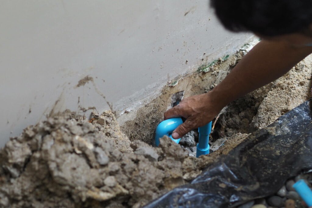 A person is working on a blue pipe, likely part of drain repairs, emerging from the ground by a wall, surrounded by earth and debris.